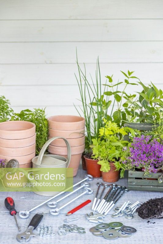 Steel rods, hooks, connectors, nuts, washers, screwdriver, spanner, watering can, pencil, terracotta pots, plants and compost laid out on a wooden surface