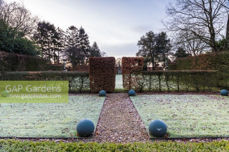View  through the frosty lawns to to the focal point at the end of the garden, as the sun rises. Painted balls form manmade structure alongside the clipped hornbeam and box hedges.