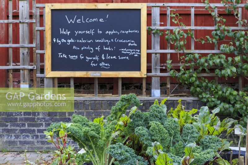 A welcome sign greets visitors entering a community garden.  Curly kale and foliage of beetroot in the foreground. Visitors are invited to pick the produce of the garden, while leaving some for others.