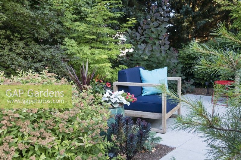 Layers of deciduous and evergreen trees, shrubs and perennials filter street view of private sitting area in front garden