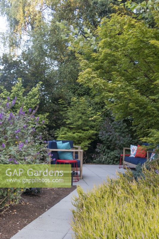 Pathway leads to secluded sitting area in front garden surrounded by layers of deciduous and evergreen trees, shrubs and perennials