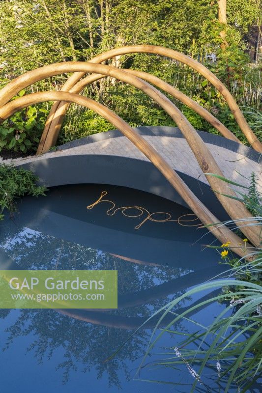 The BBC One Show and RHS Garden of Hope. Arched bridge with sculptural timber rails over tranquil pool with HOPE written in lighting in the water. Marginal planting includes marsh marigold, Caltha palustris, and white persicaria.