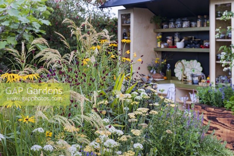 The Parsley Box Garden. Outdoor kitchen with sink and shelves. Foreground planting includes rosemary, Anemanthele lessoniana with Rudbeckia fulgida var. deamii, Achillea 'Terracotta', and garlic chives, Allium tuberosum.