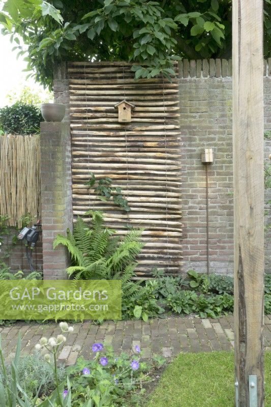 Roll of chestnut paling hung over brick wall with bird house attached, ferns and groundcover beneath.