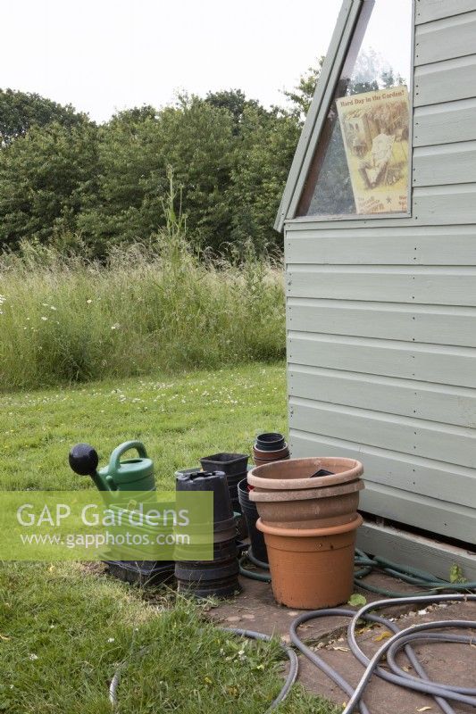 Stacked plastic pots and watering equipment outside garden shed with wildflower meadow beyond. July. Summer.