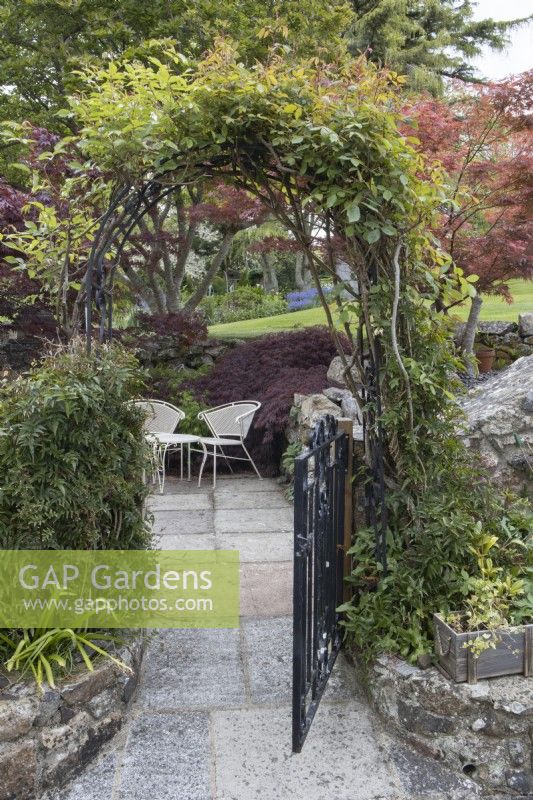 An arch and a wrought iron gate that is half open leads to a seating area with two wrought iron chairs and a table. Whitstone Farm. NGS garden, Devon. Spring. 