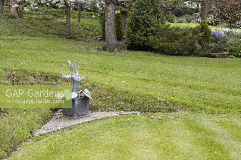 A sculpture by Matt Coe of Dingle Designs in a corner of a croquet lawn. Whitstone Farm. NGS Devon garden. Spring. 