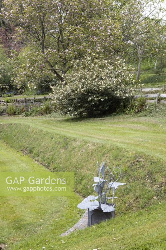 A sculpture by Matt Coe of Dingle Designs in a corner of a croquet lawn. Whitstone Farm. NGS Devon garden. Spring. 