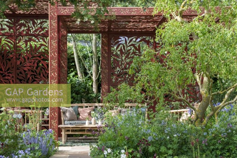 Salix x sepulcralis 'Erythroflexuosa' overhangs a border with view to a seating area with cane furniture under a laser cut pavilion - Morris  and  Co, RHS Chelsea Flower Show 2022 - Gold Medal