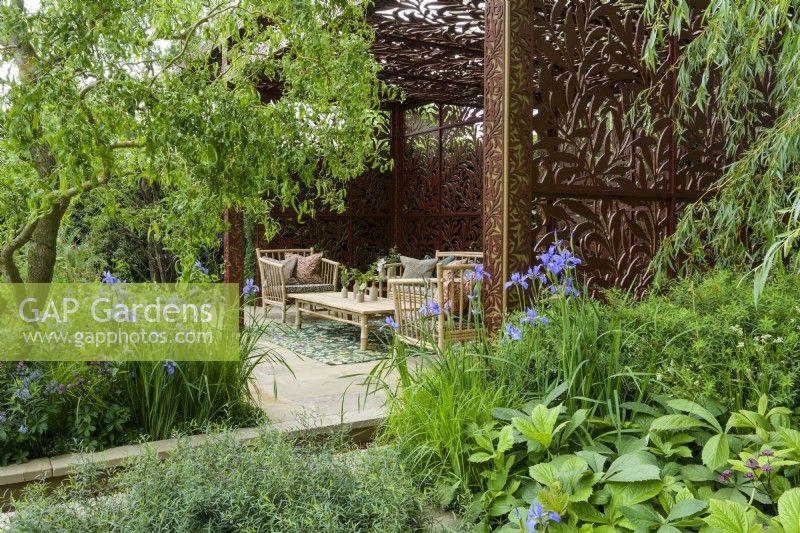 Sitting arrangement in pavillion  of metal screens  with  Morris' Willow Boughs pattern also on cushions, surrounded by herbaceous beds with blue Iris siberica, Rodgersia  and  Salix matsudana tortuosa trees, dragon's claw willow - Morris and Co. Garden