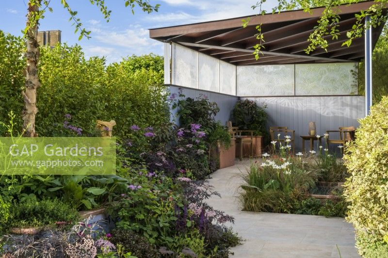 Garden with covered seating area beside a pond planted with Iris 'White Swirl' and Carex grasses with  green, purple, bronze and blue planting including Sambucus  'Black Beauty' and Betula nigra tree in raised bed  - SSAFA Sanctuary Garden. Designer: Amanda Waring