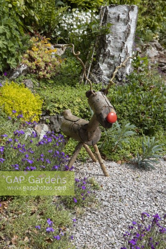 A home made rustic reindeer with red nose sits on a terrace beside purple aubretia. Briar Cottage Garden. Devon NGS garden. Spring