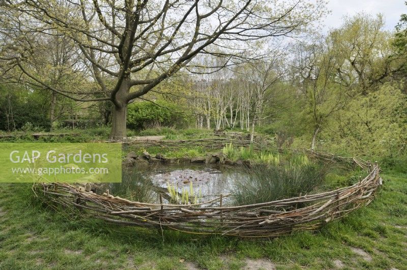 A pond in a natural setting with a low outer border fence made from twisted branches and wooden materials.