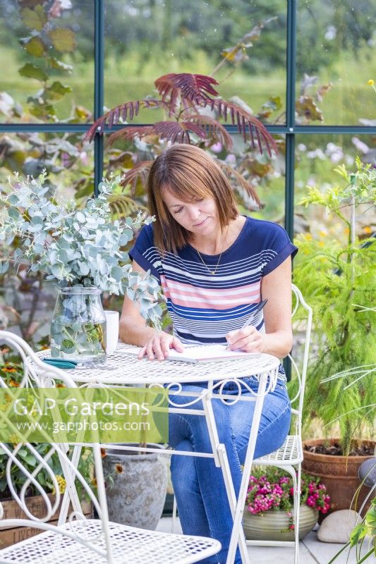 Woman sitting at table writing in a greenhouse filled with plants