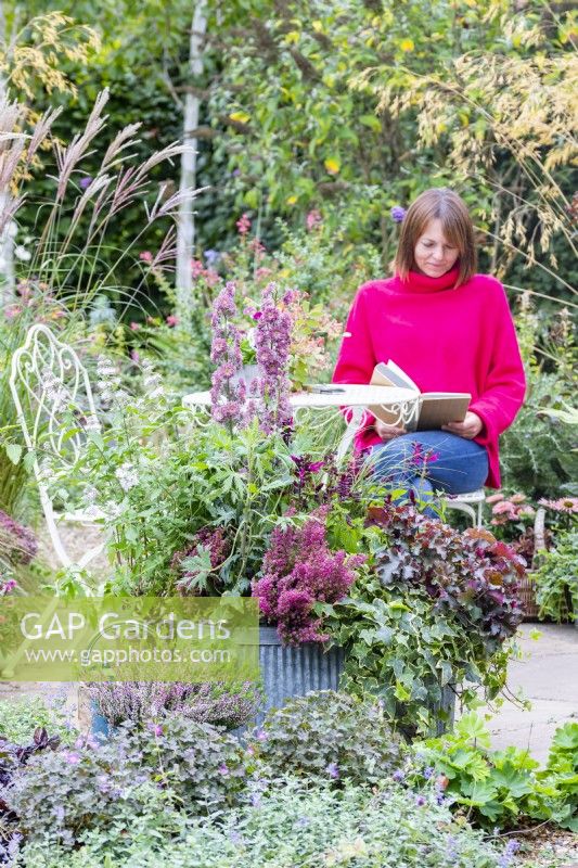 Woman sitting at a table reading on a patio behind large container planted with Delphinium 'Highlander Flamenco', Salvia 'Rockin Fuchsia', Erica gracillis, Heuchera 'Chocolate Ruffles', Stipa arundinacea and Ivy hedera