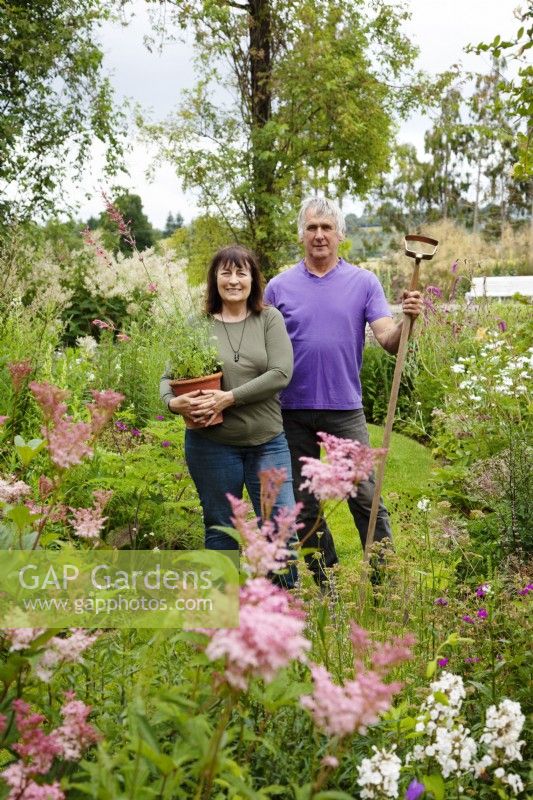 Couple standing by flower beds holding pot of Agastache 'Fleur' and a hoe.