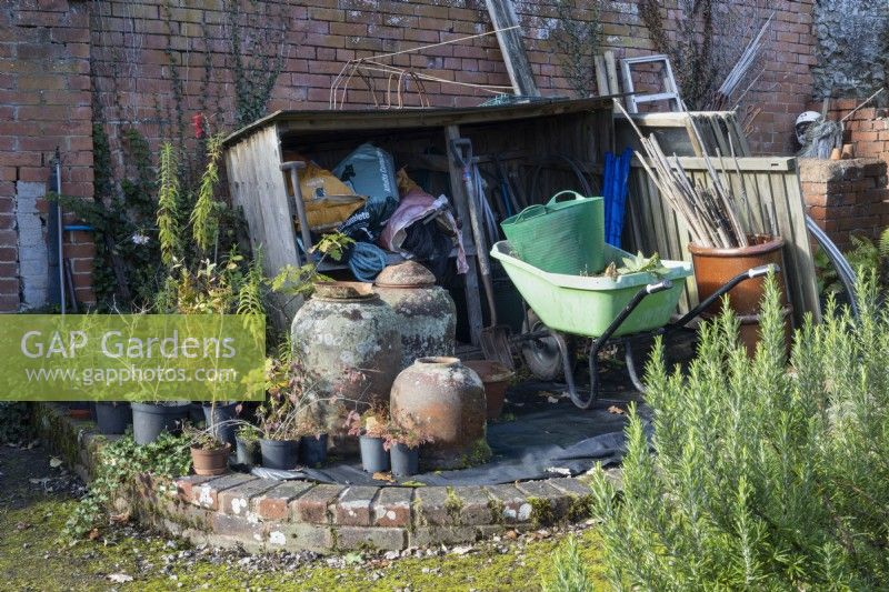 A wooden store, set against a red brick wall, holds a variety of plastic sacks with various gardening parahphanlia in front including wheelbarrow, clay cloches, plastic trug and bamboo canes. Regency House, Devon NGS garden. Autumn