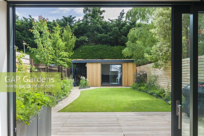 View of garden office and bicycle store at the end of a contemporary garden with lawn, stone block path, wooden decking and slatted timber fencing. June