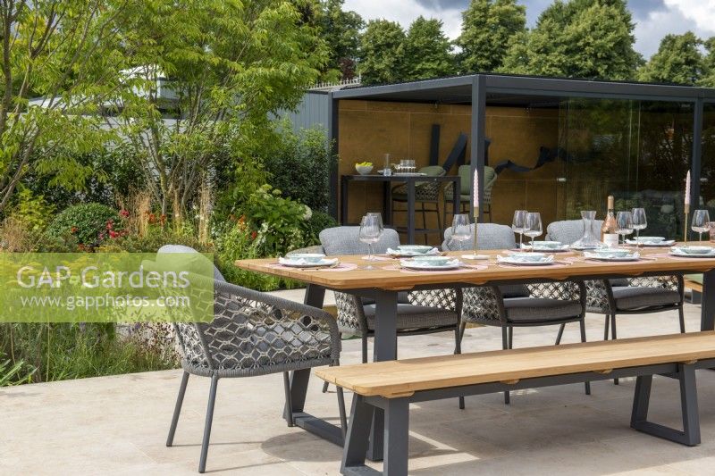 Outdoor dining table with benches and metal pergola behind - The Outdoor Living Garden, RHS Hampton Court Palace Garden Festival 2022