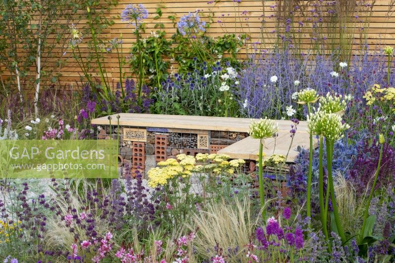 Insect hotel built into a wood and gabion bench surrounded by planting of Salvia 'Purple Rain', Gaura, Stipa tenuissima, Agapanthus 'White Heaven', Perovskia 'Blue Spire' and Achillea 'Credo' - Turfed Out Garden, RHS Hampton Court Palace Garden Festival 2022