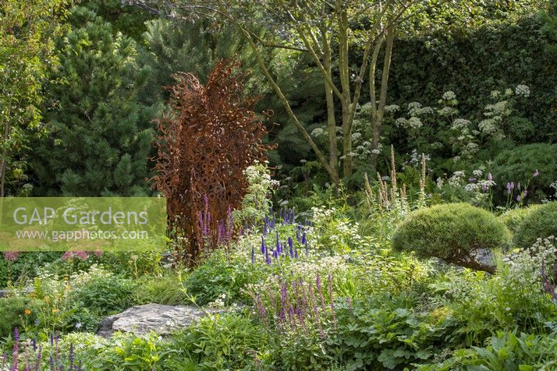 Metal sculpture by Penny Hardy surrounded by planting of Veronica longifolia and Salvia 'Caradonna', Pinus sylvestris behind - Bodmin Jail: 60 Degrees East - A Garden between Continents, RHS Chelsea Flower Show 2021
