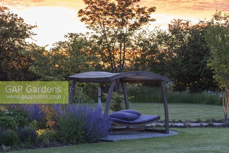 Seen after sunset, a swingseat sits on stone chippings between a lawn and a border planted with perennials.