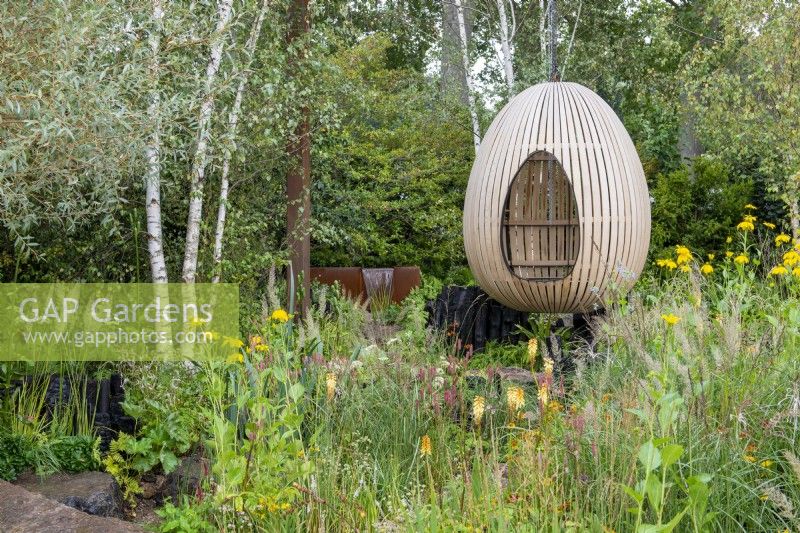 Bent oak swinging hide, suspended over naturalistic style planting, including Kniphofia, Rudbeckia 'Herbstonne' and Calamagrostis brachytrica - The Yeo Valley Organic Garden, RHS Chelsea Flower Show 2021