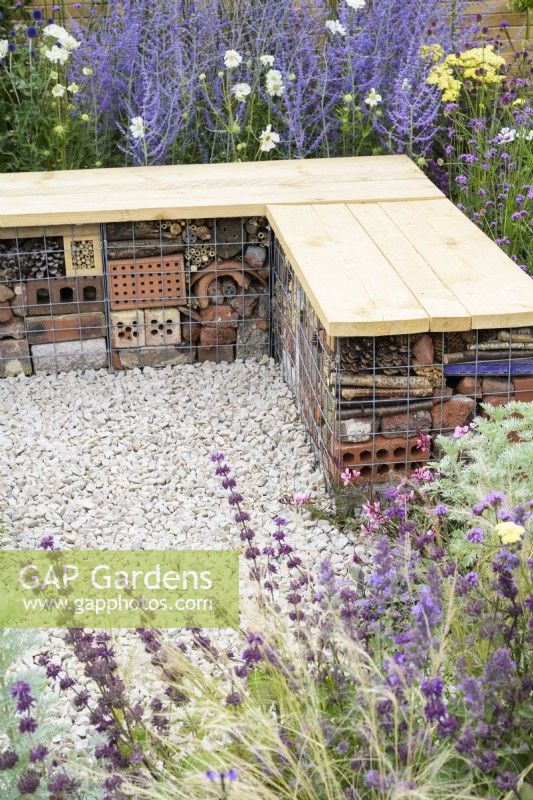Bug or insect hotel inside gabion bench with wooden top among drought-tolerant planting including Perovskia and Salvia in gravel - Turfed Out Garden, RHS Hampton Court Palace Garden Festival 2022. July. 