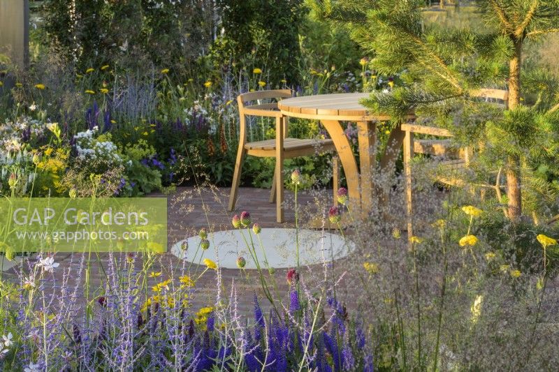 Sitting area with wooden table and chairs and planting of  Allium sphaerocephalon, Achillea 'Coronation Gold' , Agastache and Perovskia in Joy club garden - RHS Hampton Court Palace Garden Festival 2022