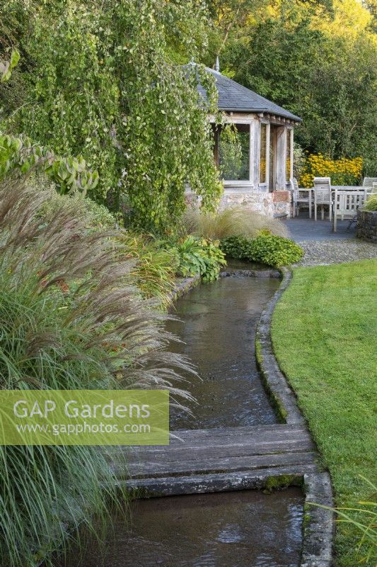 Overlooked by a summerhouse, a stream runs beside the lawn, edged in a clump of miscanthus and weeping birch, before passing beneath a planked bridge.