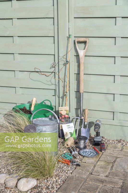 Actinidia 'Kens Red', compost, watering can, digging fork, trowel, drill, wire, tensioners, eyelet screws, pliers, tape measure, pencil, scissors, secateurs and rootgrow laid out on the ground