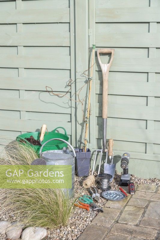 Actinidia 'Kens Red', compost, watering can, digging fork, trowel, drill, wire, tensioners, eyelet screws, pliers, tape measure, pencil, scissors and secateurs laid out on the ground
