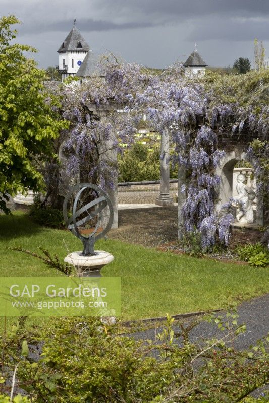 An armillary sundial on pedestal with wisteria covered pergola behind. Trago Mills show gardens, Devon, UK. May. Spring