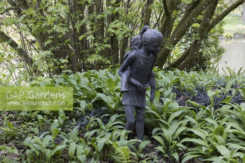 A statue of a girl carrying a small child, piggyback amongst spring growth of wild garlic. Marwood Hill gardens, Devon. Spring. Devon. 