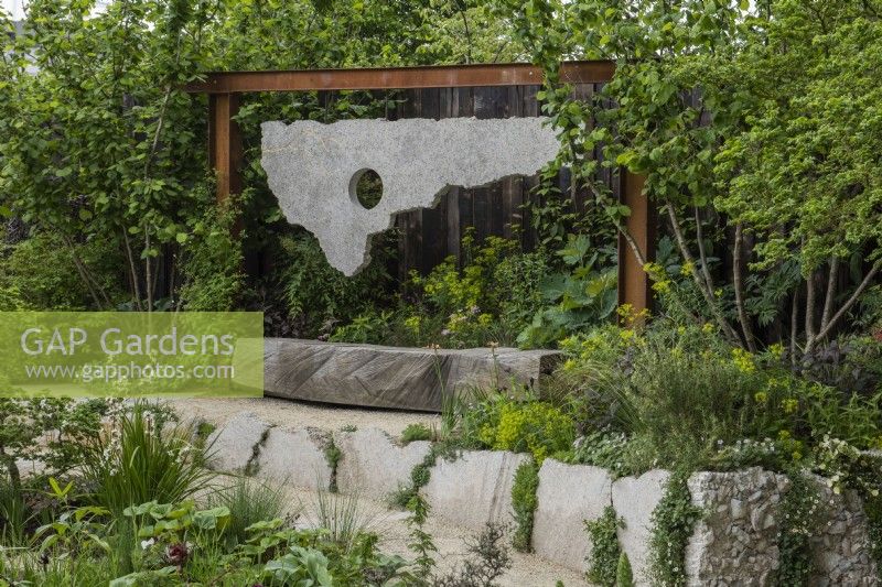 A wooden bench is backed by a sculpture made from salvaged materials, softened by perennials and hazel trees, Corylus avellana, to each side.