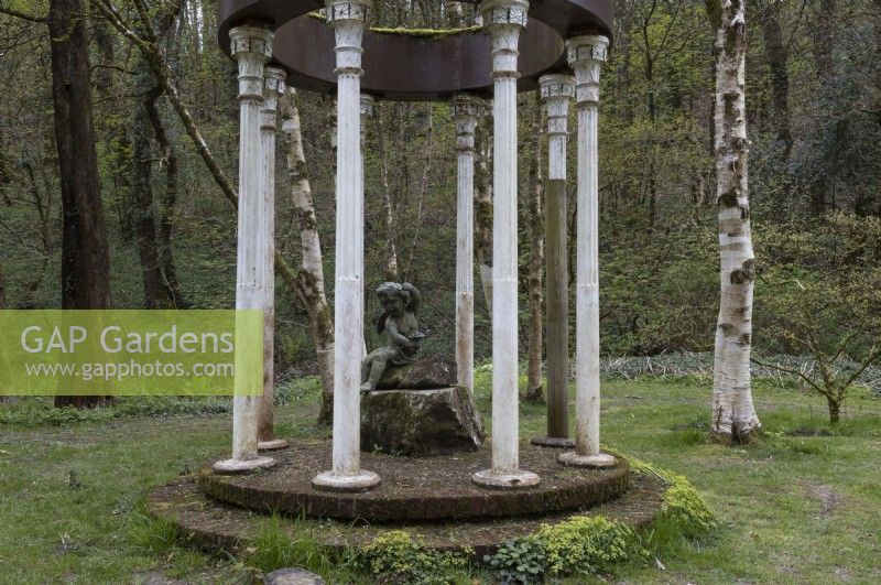 The Folly, a structure of columns with a tree stump in the centre and a statue of a cherub on top in a wooded garden. Marwood Hill Gardens. Spring. Devon. May. 