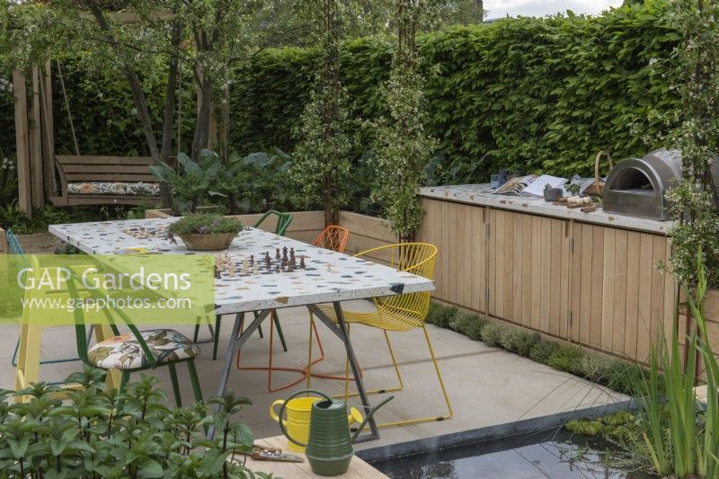 A community garden enclosed in hornbeam hedges, separated by raised beds of vegetables into a dining area with work area and storage, and a quiet space with swingseat shaded by trees.