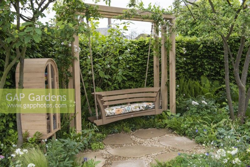 A quiet space with swingseat shaded by trees and edged in borders of ferns and white flowers. A wooden cabinet houses books, seeds and cushions.