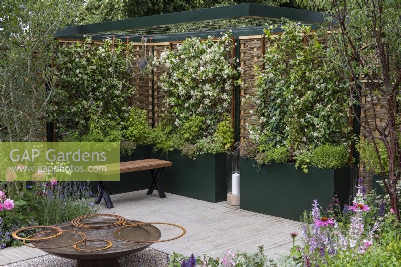 A formal courtyard is enclosed in willow and hazel fencing screens, supporting fragrant star jasmine, Trachelospermum jasminoides, which grows in tall planters with herbs. A water feature is crafted from reclaimed aluminium. Borders contain roses and herbaceous perennials.
