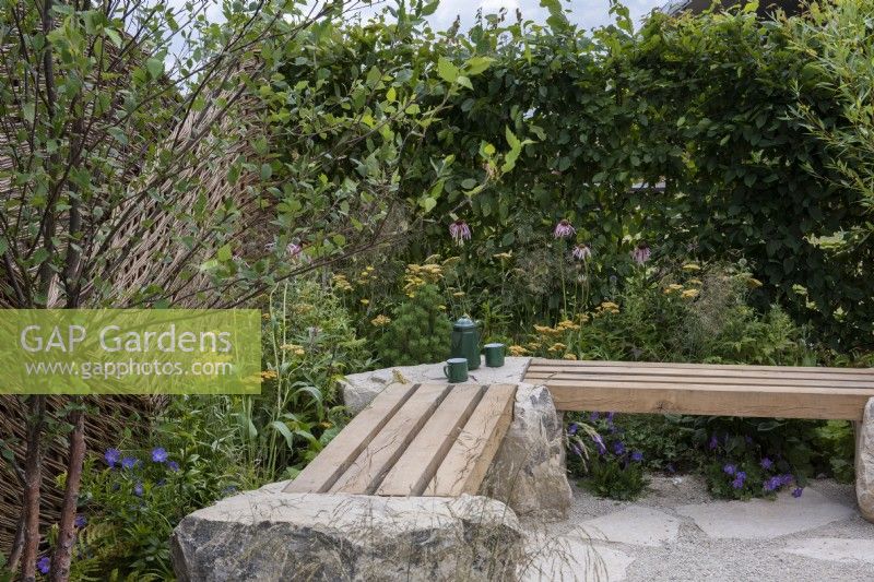 The corner of a small courtyard is enclosed in a hornbeam hedge and willow fence, and edged in flowering perennials. A  bench is crafted from reclaimed oak sleepers, and anchored in Purbeck limestone boulders.