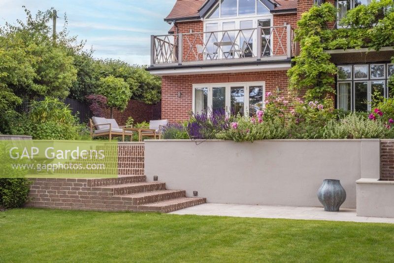 View of brick steps and white rendered retaining wall leading up to raised terrace and house with mixed borders and furniture 