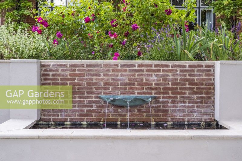 Verdigris basin fountain on brick wall of raised rendered water feature with terrace border with roses above.