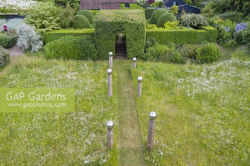 View over meadow with formal avenue of cut grass lined with wooden posts topped with stainless steel globes. Border of Hydrangeas with hedge of Box and large clipped hornbeam entrance June. Summer. Image taken with drone. 
