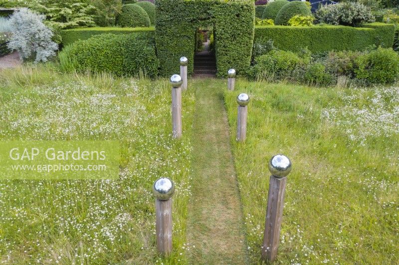 View over meadow with formal avenue of cut grass lined with wooden posts topped with stainless steel globes. Hedge of Box and large clipped hornbeam entrance June. Summer. Image taken with drone. 