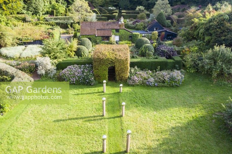 Meadow that has been cut with avenue of columns of wood capped with stainless steel globes and backed by border of Hydrangeas and a clipped tunnel of Hornbeam. View to the house beyond. September. Image taken with drone. 