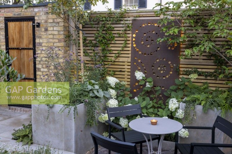 Courtyard garden at dusk with raised borders and contemporary wood boundary fence with metal screen and lighting