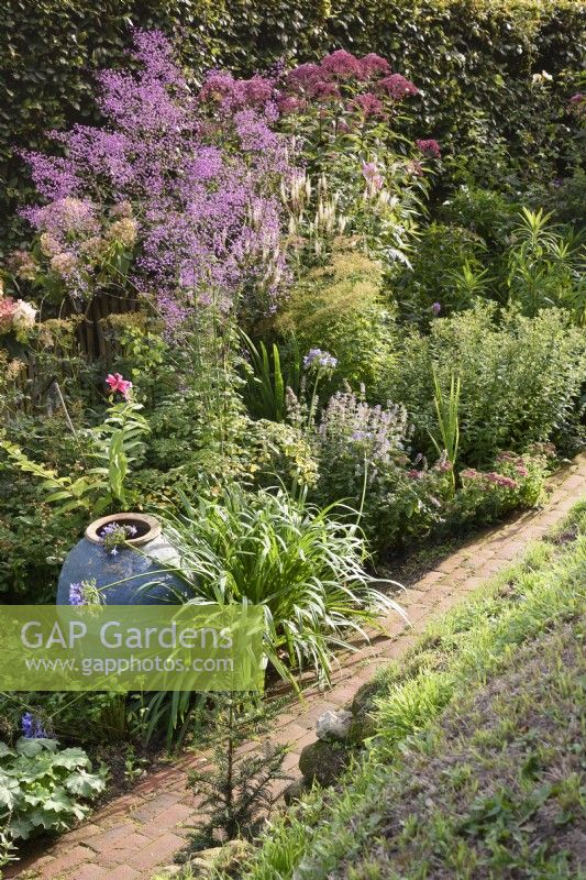 View down onto a brick path in August, beyond a perennial border with ornamental empty urn