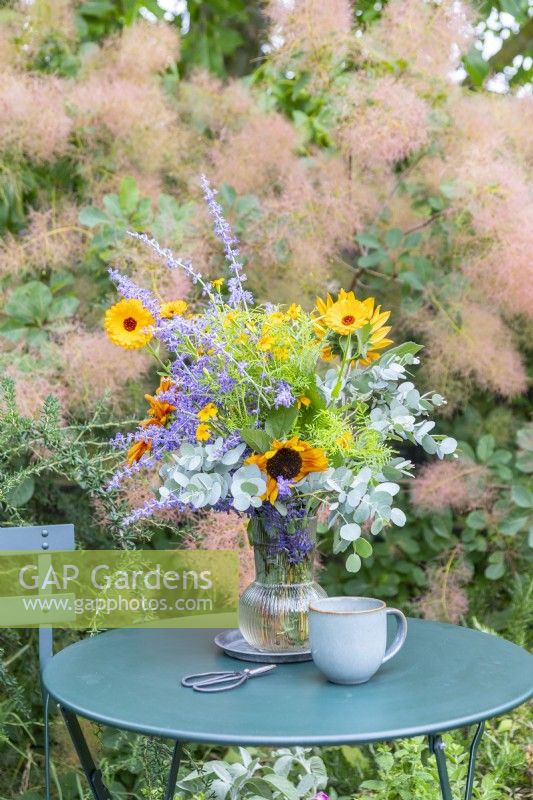 Bouquet containing Helianthus - Sunflowers, Eucalyptus sprigs, Tagetes 'Lemon Gem', Perovskia and Calendula 'Indian Prince' in glass vase on small garden table with snips and a mug