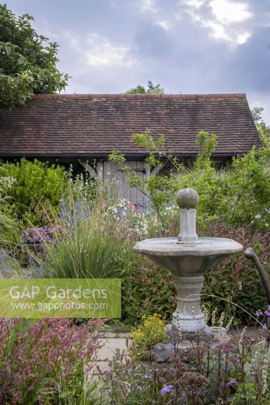 Small courtyard garden filled with late summer planting, with framed oak building behind and water feature, bird bath in foreground