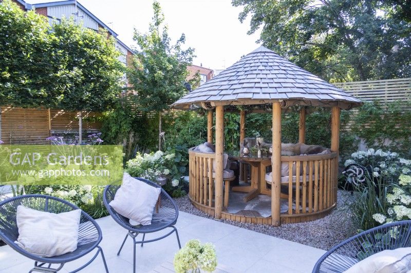 Round tile roofed wooden gazebo with seating and table inside next to stone tile patio surrounded by mixed planting